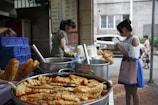 A street food stall displays a variety of fried pastries and food items in metal trays, with large cooking pots in the background. Two women are seen, one purchasing food and the other working at the stall. Beverages in plastic cups are also visible on the counter.