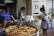 A street food stall displays a variety of fried pastries and food items in metal trays, with large cooking pots in the background. Two women are seen, one purchasing food and the other working at the stall. Beverages in plastic cups are also visible on the counter.