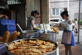 A street food stall displays a variety of fried pastries and food items in metal trays, with large cooking pots in the background. Two women are seen, one purchasing food and the other working at the stall. Beverages in plastic cups are also visible on the counter.