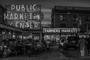 A bustling urban scene featuring a public market with neon signs reading 'Public Market Center' and 'Farmers Market'. The setting is evening, as indicated by the illuminated signs and dark sky. People are walking around, and several parked cars are visible, creating an atmosphere of lively commerce.