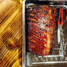 A tray with grilled ribs glazed in barbecue sauce, garnished with sesame seeds. Scissors are placed next to the ribs, and a glass filled with a brown liquid sits on the wooden table.