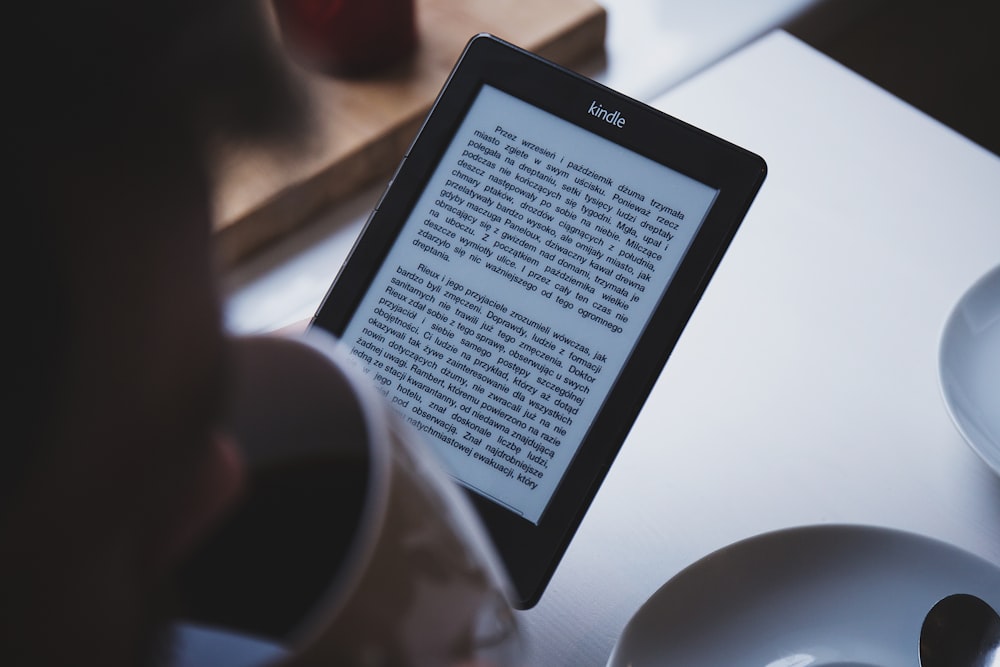 A person reading a book on a Kindle while drinking coffee