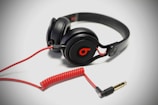 A pair of black over-ear headphones with a red cable and connector, featuring a distinctive logo on the ear cups. The cable is coiled and extends to a gold-plated plug.