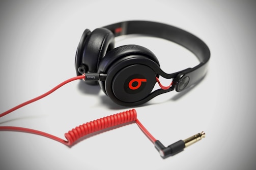 A pair of black over-ear headphones with a red cable and connector, featuring a distinctive logo on the ear cups. The cable is coiled and extends to a gold-plated plug.