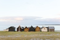 Several rustic huts made of wood and stone stand in a row on a grassy shore beside a calm sea. The sky is partly cloudy, suggesting a serene and peaceful seaside setting.