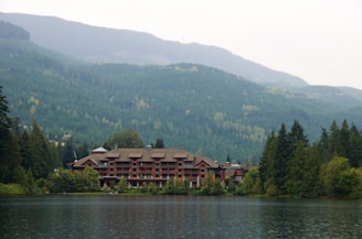A large lodge-style building sits nestled among lush green forests and overlooks a calm lake. The backdrop features rolling mountains covered in dense forests under a slightly overcast sky. The serene setting conveys a deep sense of tranquility and natural beauty.