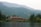 A large lodge-style building sits nestled among lush green forests and overlooks a calm lake. The backdrop features rolling mountains covered in dense forests under a slightly overcast sky. The serene setting conveys a deep sense of tranquility and natural beauty.