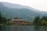 A large lodge-style building sits nestled among lush green forests and overlooks a calm lake. The backdrop features rolling mountains covered in dense forests under a slightly overcast sky. The serene setting conveys a deep sense of tranquility and natural beauty.