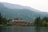 A large lodge-style building sits nestled among lush green forests and overlooks a calm lake. The backdrop features rolling mountains covered in dense forests under a slightly overcast sky. The serene setting conveys a deep sense of tranquility and natural beauty.