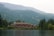A large lodge-style building sits nestled among lush green forests and overlooks a calm lake. The backdrop features rolling mountains covered in dense forests under a slightly overcast sky. The serene setting conveys a deep sense of tranquility and natural beauty.