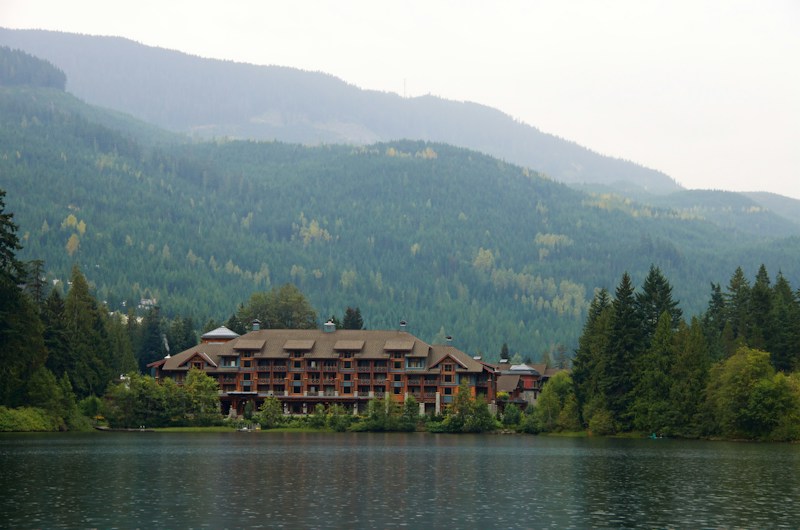 A large lodge-style building sits nestled among lush green forests and overlooks a calm lake. The backdrop features rolling mountains covered in dense forests under a slightly overcast sky. The serene setting conveys a deep sense of tranquility and natural beauty.