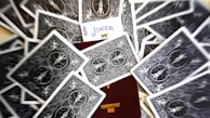 A scattered deck of playing cards surrounds a partially visible passport. The card with the Joker face is prominently displayed, positioned above the passport.