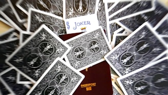 A scattered deck of playing cards surrounds a partially visible passport. The card with the Joker face is prominently displayed, positioned above the passport.