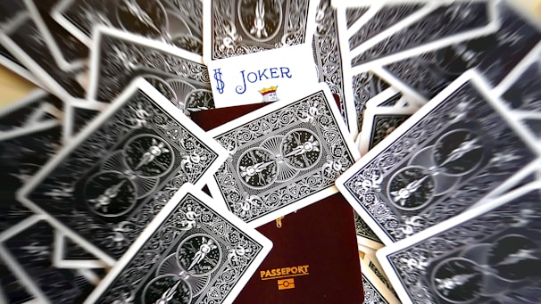A scattered deck of playing cards surrounds a partially visible passport. The card with the Joker face is prominently displayed, positioned above the passport.