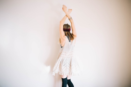 A person with long hair stands facing a plain wall, arms raised above their head, wearing a light, flowing dress and knee-high socks. The individual has a headband and bracelets, creating a bohemian or carefree style.