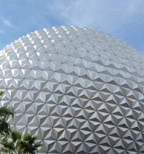 A large geodesic sphere with a complex triangular panel pattern against a clear blue sky, with lush green palm leaves in the corner.