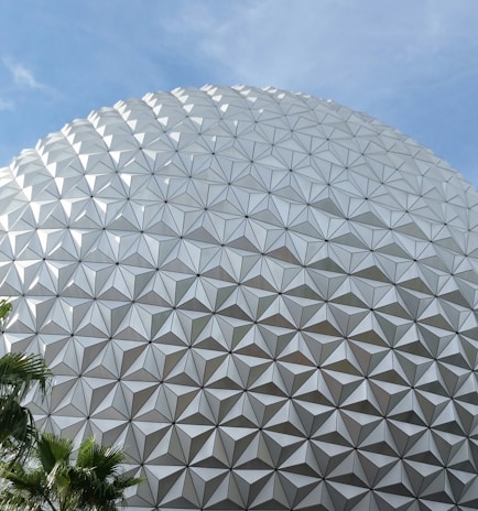 A large geodesic sphere with a complex triangular panel pattern against a clear blue sky, with lush green palm leaves in the corner.