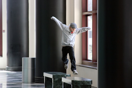 A person wearing a gray hoodie and cap is jumping from one marble platform to another in a corridor with large dark pillars and tall windows. The person appears to be engaged in a parkour or playful activity.