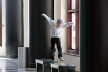 A person wearing a gray hoodie and cap is jumping from one marble platform to another in a corridor with large dark pillars and tall windows. The person appears to be engaged in a parkour or playful activity.