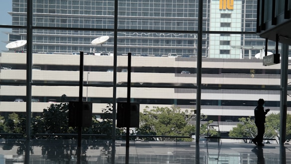 The scene includes a silhouette of a person inside a building with large windows providing a view of the outside. Beyond the windows, there is a parking structure with multiple levels and a set of satellite dishes on top. In the background, a modern office building features reflective windows. The interior has a polished floor, and there are trees visible outside.