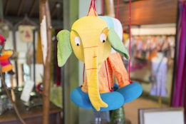 A colorful, fabric elephant toy is suspended by red strings. It features a bright yellow face, large green ears, a long nose, and blue padded feet. The toy has a whimsical design with different patterns, including a pink and yellow striped body. Behind it, a blurred background displays various colorful textiles and items, suggesting a shop setting.