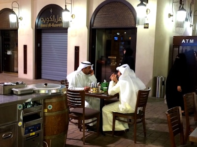Two men dressed in traditional white garments are seated at a small round table outdoors, possibly having a conversation. A tea pot and cups are on the table. The setting appears to be in a cafe or a small restaurant, with wooden chairs and a barrel-shaped fixture. There is an ATM in the background, along with a closed shop front labeled 'Amal Al-Kuwait'. The area is illuminated by warm lighting sconces attached to the walls.
