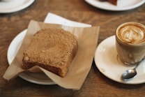 A piece of thick, crumbly toast is placed on a piece of brown parchment paper on a white plate. Next to it, a cappuccino or latte in a clear glass sits on a matching saucer, accompanied by a small spoon. The table beneath is made of a rustic wood.