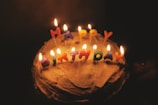 A cake with lit candles spelling out 'HAPPY BIRTHDAY' placed on top, surrounded by a dark background that emphasizes the warm glow of the flames.