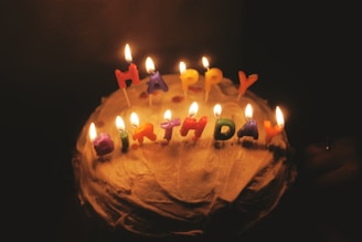 A cake with lit candles spelling out 'HAPPY BIRTHDAY' placed on top, surrounded by a dark background that emphasizes the warm glow of the flames.