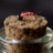 A close-up view of a fishing fly placed on the top of a cork stopper. The fly has a vibrant red body with some black and metallic elements, likely representing a small insect or creature. The cork stopper is light brown with a rough texture, set atop a glass container.