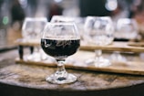 A glass of dark beer sits on a wooden barrel, with blurred glassware and wooden paddles in the background, creating a rustic and cozy atmosphere.