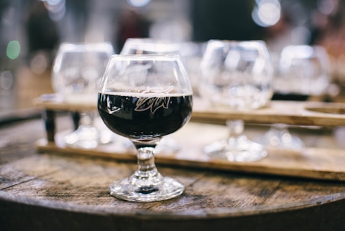 A glass of dark beer sits on a wooden barrel, with blurred glassware and wooden paddles in the background, creating a rustic and cozy atmosphere.
