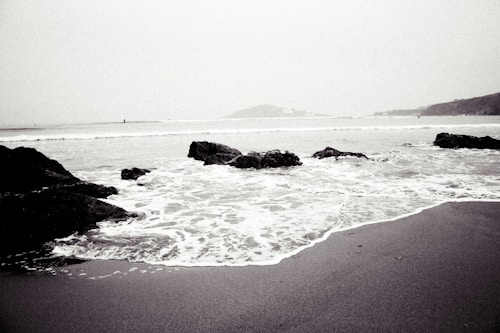 A grayscale image featuring a serene beach with gentle waves lapping against the shore. Large rocks are scattered along the sand, extending into the water. The horizon is barely visible under a misty sky, adding a sense of calm and solitude to the scene.