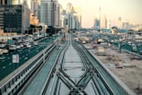 Urban landscape with high-rise buildings and a busy highway. Train tracks in the foreground lead towards the city, surrounded by construction areas and numerous vehicles. The skyline includes modern skyscrapers and a clear sky.