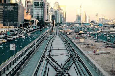 Urban landscape with high-rise buildings and a busy highway. Train tracks in the foreground lead towards the city, surrounded by construction areas and numerous vehicles. The skyline includes modern skyscrapers and a clear sky.