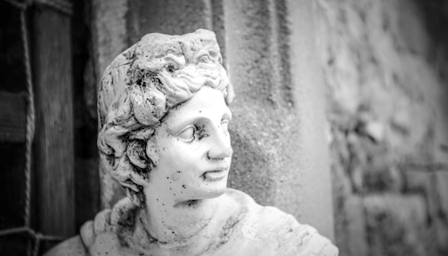 A classical stone sculpture of a person with detailed hair and facial features, positioned against a textured wall. The statue appears weathered, with visible marks and discoloration.