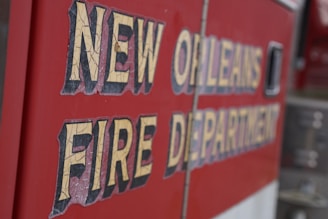 Text on the side of a vehicle reads 'New Orleans Fire Department' in bold, weathered lettering. The background is a vibrant red, typical of emergency service vehicles.