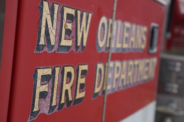 Text on the side of a vehicle reads 'New Orleans Fire Department' in bold, weathered lettering. The background is a vibrant red, typical of emergency service vehicles.