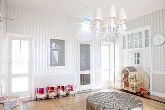 An airy and well-lit children's playroom featuring a soft color palette. The room includes a large round ottoman with a metallic finish in the center, toy carts lined up against the wall, and a wooden dollhouse with various miniature furniture. A modern chandelier with multiple white lampshades hangs from the ceiling. The walls are decorated with vertical stripes and include several white-framed glass doors and windows. A clock hangs on the wall near the ceiling.