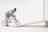 A doll riding a miniature red bicycle is positioned on a wooden ramp. The doll is dressed in a blue shirt and beige pants, and the setting is a simplistic, minimalist background that highlights the action of the scene.