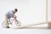 A doll riding a miniature red bicycle is positioned on a wooden ramp. The doll is dressed in a blue shirt and beige pants, and the setting is a simplistic, minimalist background that highlights the action of the scene.