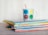 A stack of colorful children's books is placed on a wooden surface. On top of the books sits a playful mug featuring various cheerful faces in different colors, with a red straw sticking out. The colors of the book spines vary, adding a vibrant and lively touch to the scene.