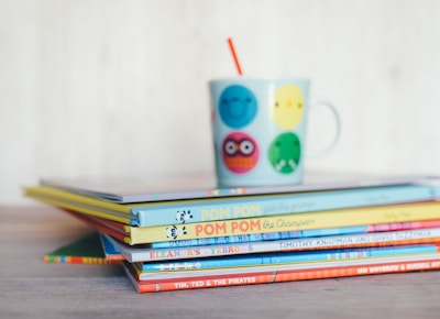 A stack of colorful children's books is placed on a wooden surface. On top of the books sits a playful mug featuring various cheerful faces in different colors, with a red straw sticking out. The colors of the book spines vary, adding a vibrant and lively touch to the scene.