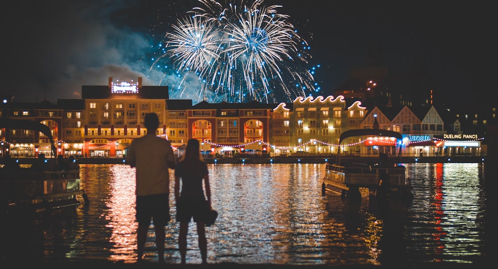 Dos personas de pie junto a un cuerpo de agua mirando fuegos artificiales