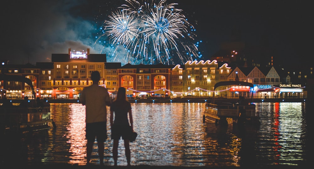 two people standing beside body of water watching fireworks