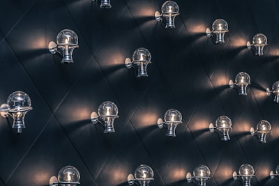 Numerous wall-mounted light fixtures arranged in a geometric pattern against a dark, diamond-patterned wall. Each fixture emits a soft, warm glow, illuminating the space subtly.