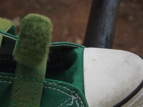 A close-up view of a green sneaker with a white toe cap, showcasing its fabric texture and stitching details. The shoe's tongue is visible, made of a fuzzy green material. The focus is on the upper part of the shoe, displaying signs of wear.