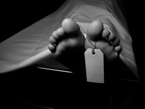 Bare feet are visible under a white sheet, with a tag attached to one of the toes, suggesting a morgue or autopsy setting. The lighting is dim, emphasizing a somber atmosphere.