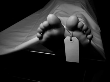 Bare feet are visible under a white sheet, with a tag attached to one of the toes, suggesting a morgue or autopsy setting. The lighting is dim, emphasizing a somber atmosphere.