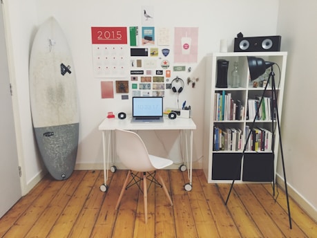 A neatly organized home office or study space featuring a small white desk with a laptop showing the time, a white chair, and various stationery items. On the wall, there is a calendar and an assortment of pictures and notes. A surfboard stands leaning against the wall on the left side. A white bookshelf on the right holds a collection of books, a pair of speakers on top, and a floor lamp illuminating the area.
